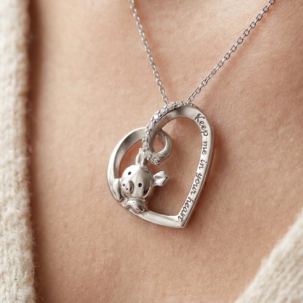 Silver heart pendant with a small pig inside the heart, a row of tiny stones on the top loop and the words Keep me in your heart engraved on the edge, shown on a fine chain (styled lifestyle photo, pendant only, chain sold separately)